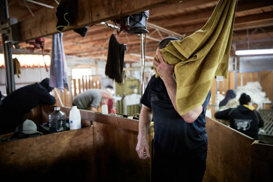 Adult male wiping face with towel inside industrial structure with wooden walls. Other people and supplies are visible in background.