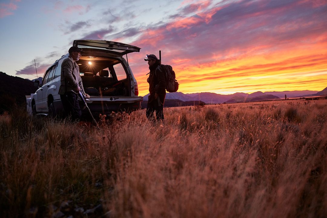 Two adult males stand near an SUV with its hatch open in a field at sunset with mountains visible in the background.