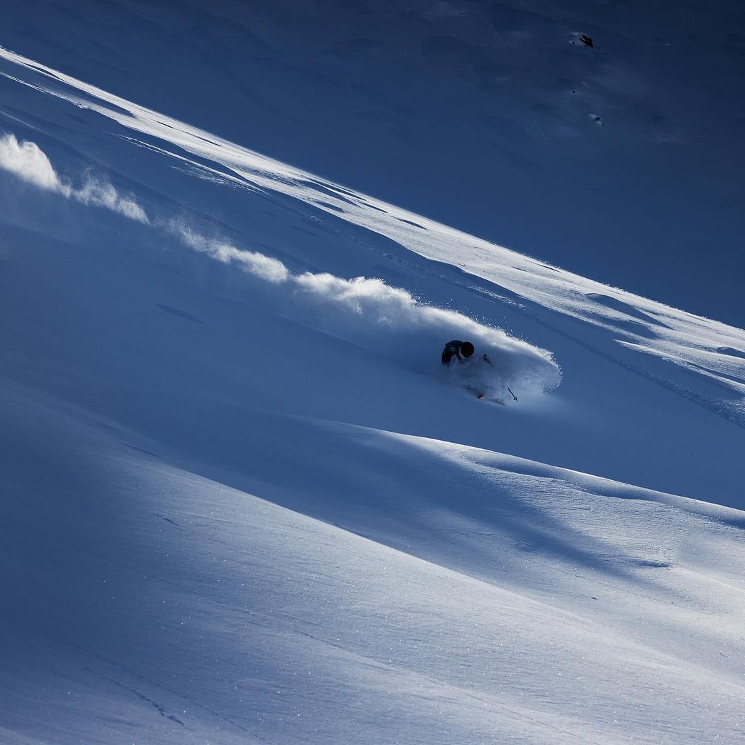 Person skiing down a snow-covered mountain on a sunny day, creating a cloud of snow powder in a dynamic, outdoor action shot.