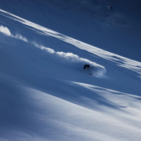 Person skiing down a snow-covered mountain on a sunny day, creating a cloud of snow powder in a dynamic, outdoor action shot.