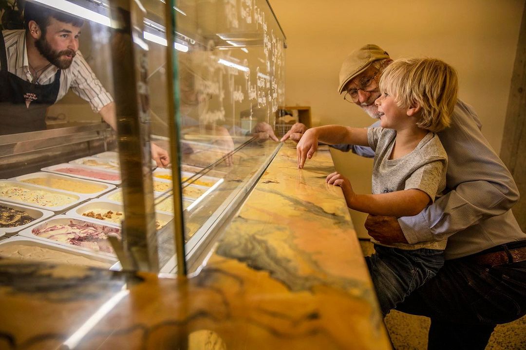 A young child and an older adult look into a glass display case filled with various flavors of ice cream, served by a bearded server.