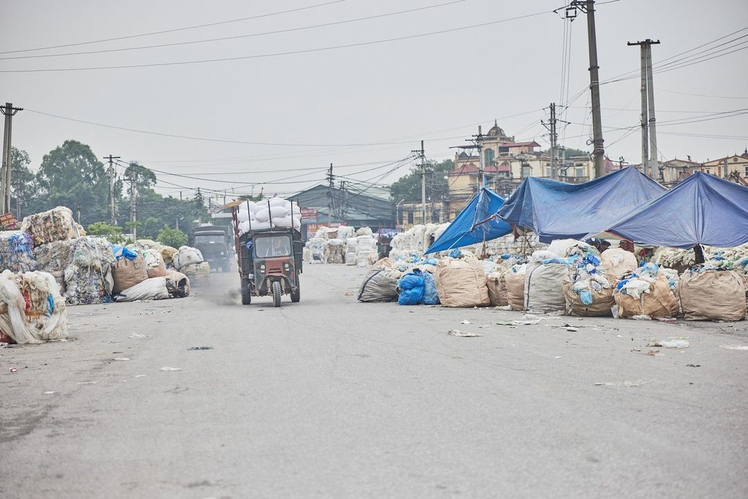 Tricycle laden with white sacks drives down road, with piles of garbage bags on either side, under an overcast sky.