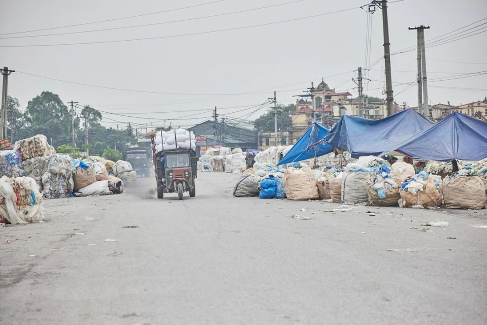 Tricycle laden with white sacks drives down road, with piles of garbage bags on either side, under an overcast sky.