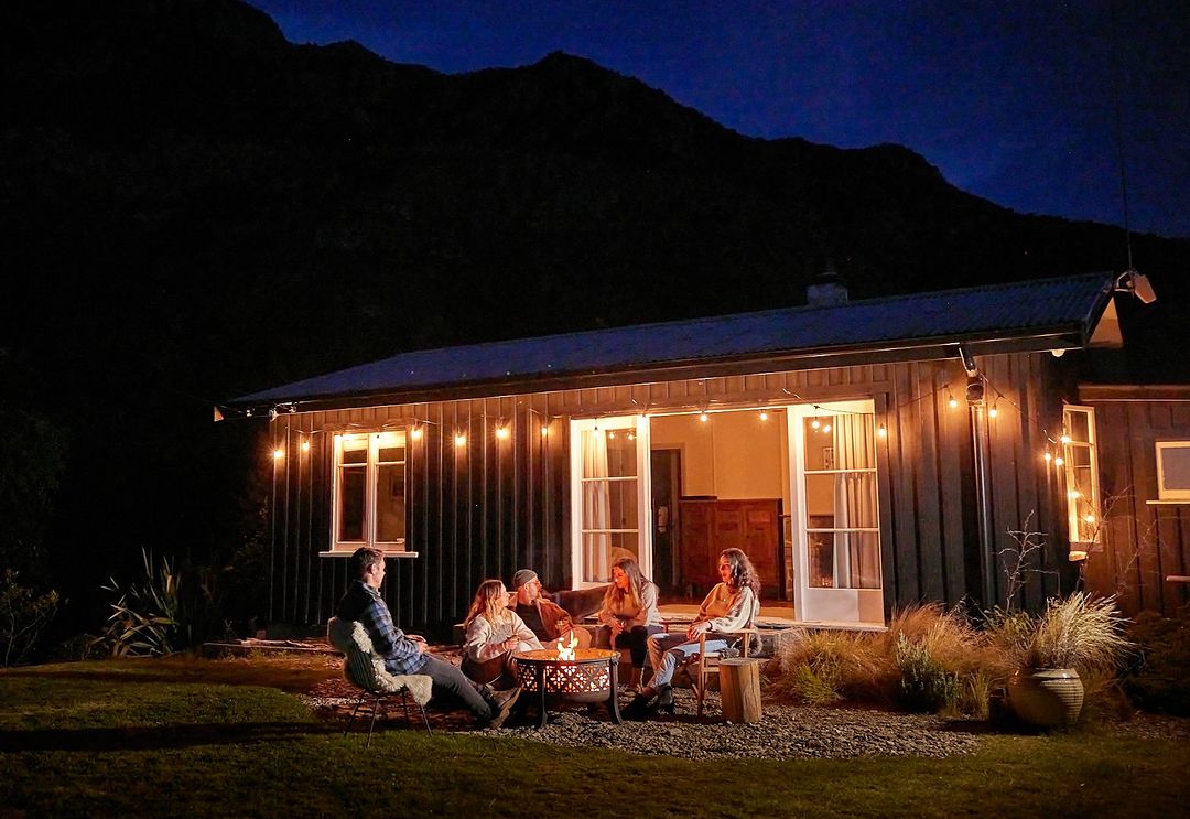 Five people gathered around a fire pit outside a cozy cottage at night, with mountains visible in the background under a starlit sky.