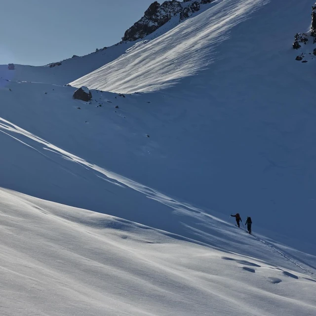 Two people ski touring up a snowy mountain slope under a clear sky, creating tracks in the snow with a rocky mountain ridge visible.