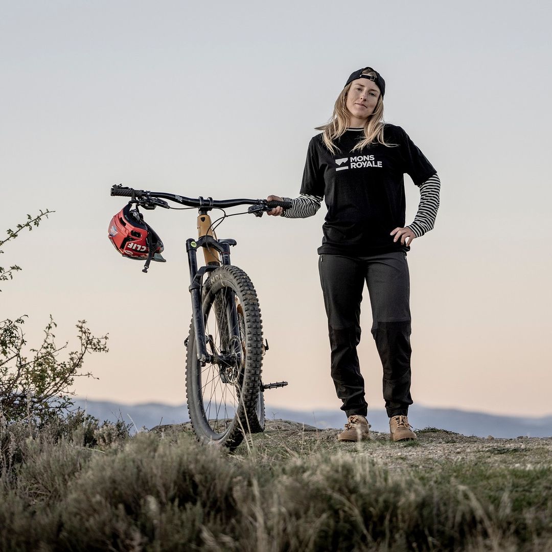 Female mountain biker standing with her bicycle wearing a Mons Royale T-shirt with a helmet hanging from the handlebar.