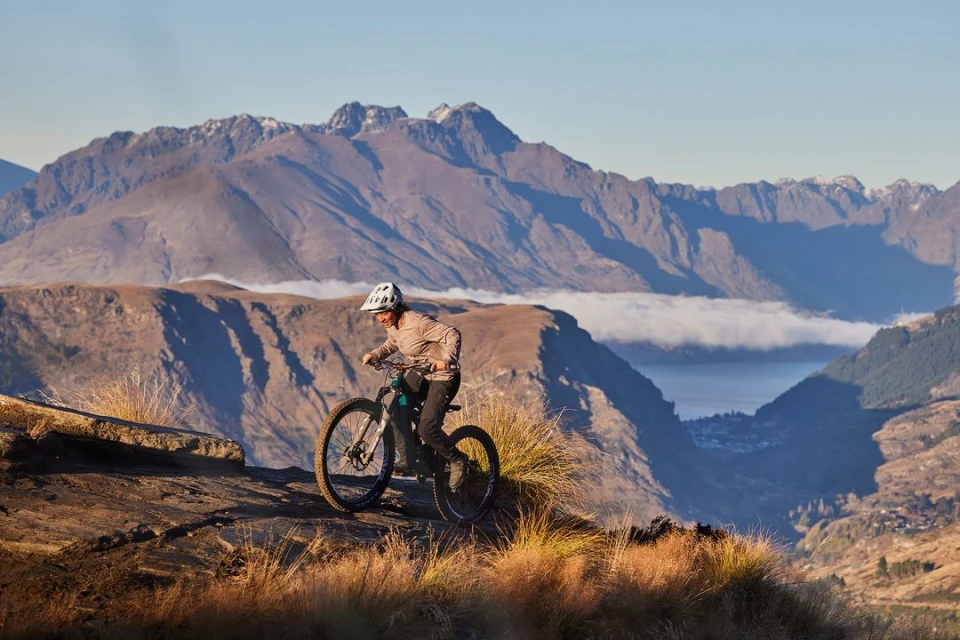 Woman mountain biking on a rocky trail with a scenic view of mountains and a lake in the background, wearing helmet, athletic clothing.