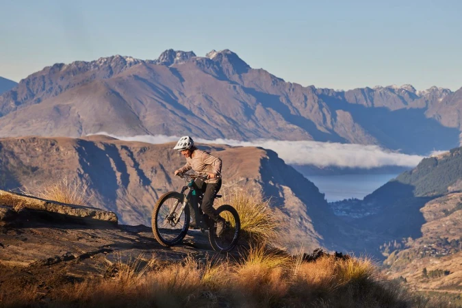 Woman mountain biking on a rocky trail with a scenic view of mountains and a lake in the background, wearing helmet, athletic clothing.