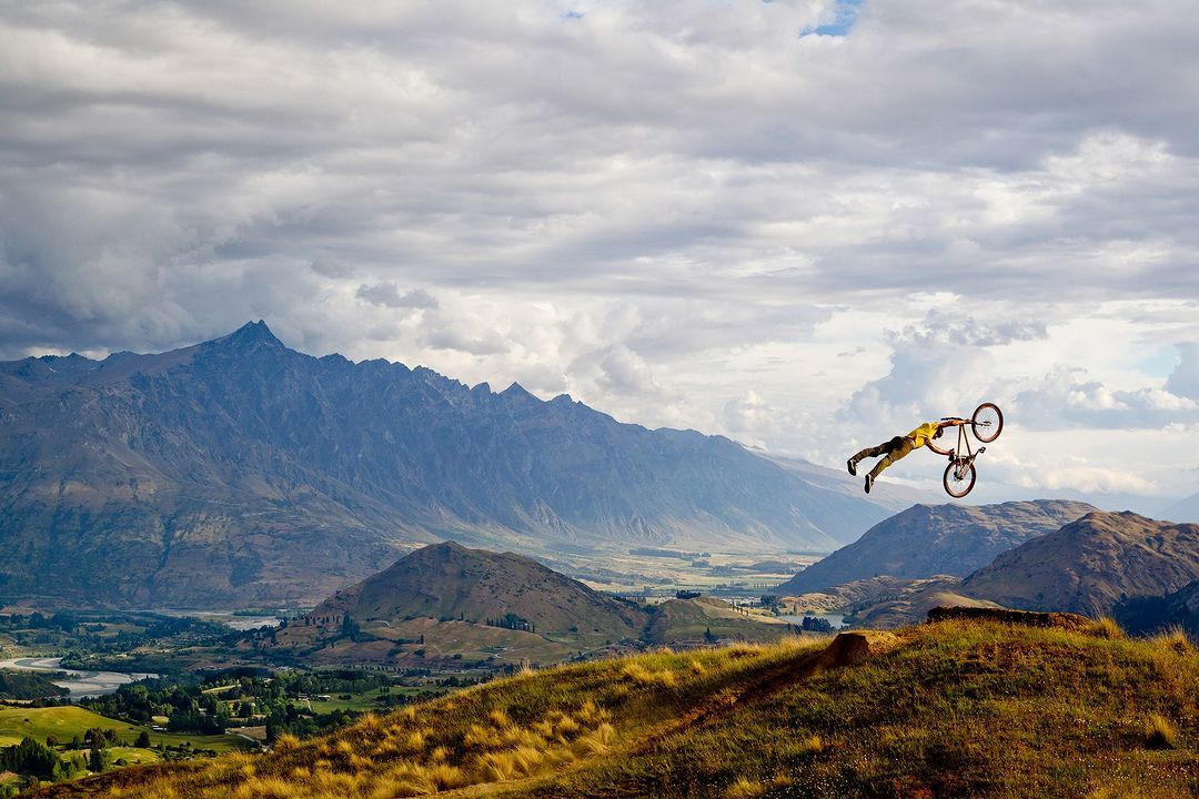 Person performing a bike trick on a mountain bike in mid-air over a hillside landscape with mountains and a cloudy sky in the background.