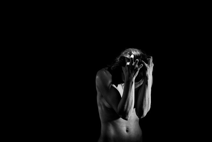 Black and white portrait of an adult woman with a partially nude torso and arms raised to her head against a dark background.