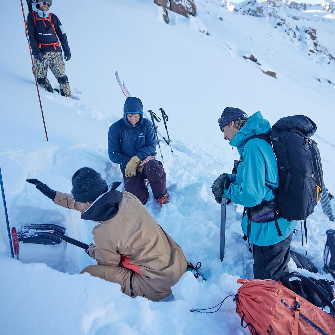 Four people in winter gear are digging a snow pit on a sunny, snowy mountain, with backpacks, skis, and shovels visible.