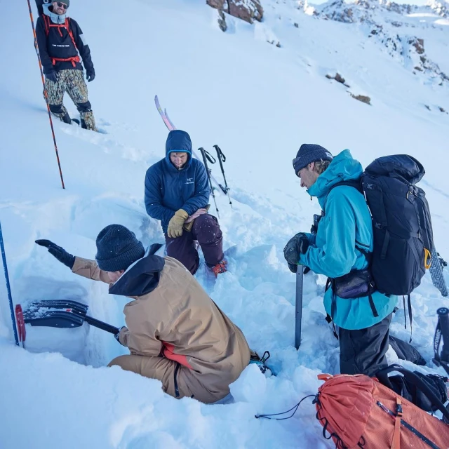 Four people in winter gear are digging a snow pit on a sunny, snowy mountain, with backpacks, skis, and shovels visible.