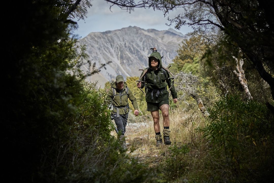 Two hikers in outdoors clothing walk a grassy trail in a lush forest toward mountain peaks in New Zealand in natural daylight.