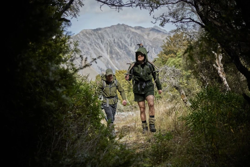 Two hikers in outdoors clothing walk a grassy trail in a lush forest toward mountain peaks in New Zealand in natural daylight.