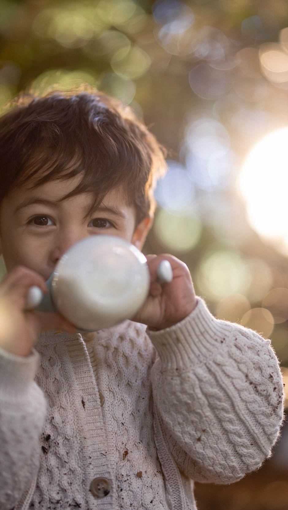 Child drinks from a bottle outdoors in a close-up shot; they are wearing a cable knit sweater and there are blurred trees behind.