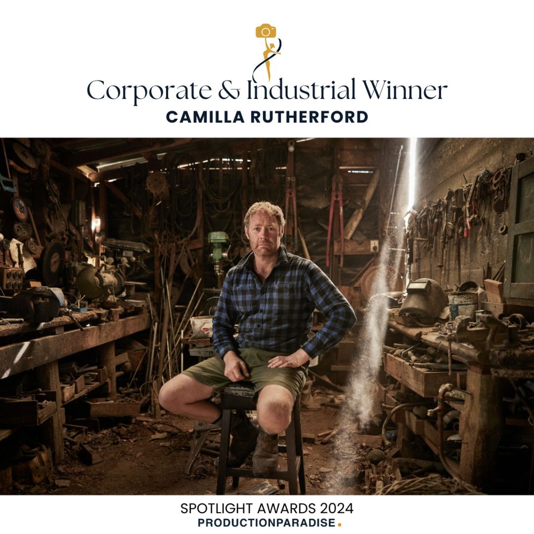 A man poses in an industrial workshop, seated on a stool. Light streams through the cluttered space with tools, equipment, and rustic charm.