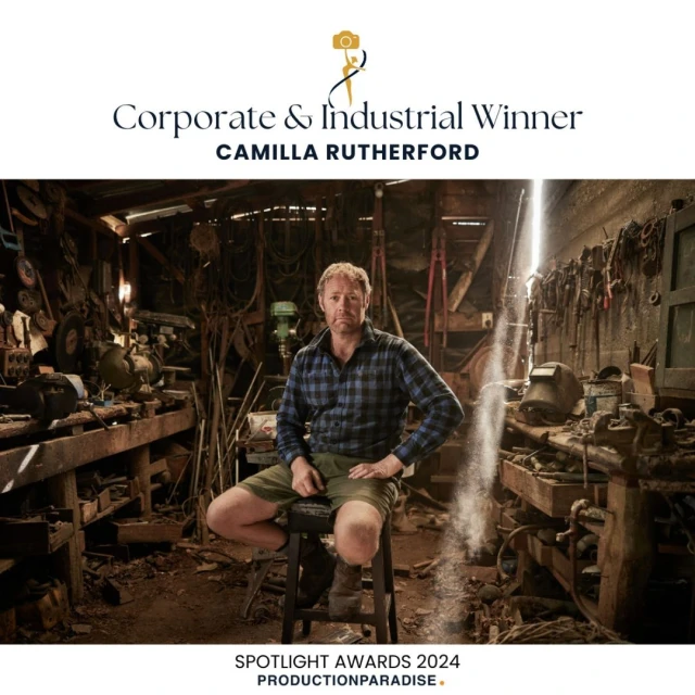 A man poses in an industrial workshop, seated on a stool. Light streams through the cluttered space with tools, equipment, and rustic charm.