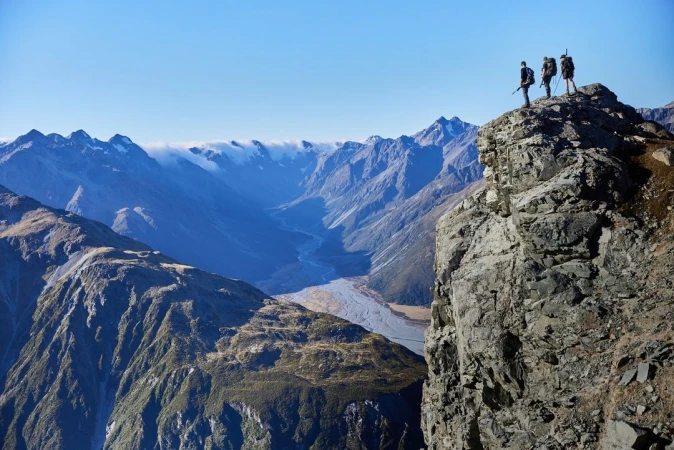 Three people with backpacks stand on a rocky cliff overlooking a mountainous landscape under a clear blue sky, sunny daytime.