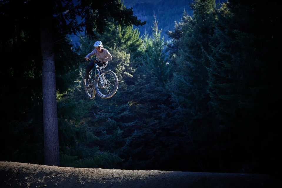 An adult rides a mountain bike, soaring through the air in a forest setting, demonstrating extreme sports and outdoor activities.
