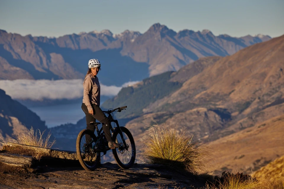 Adult woman wearing a helmet and outdoor sportswear poses with a mountain bike on a mountain top with valley and mountain range backdrop.