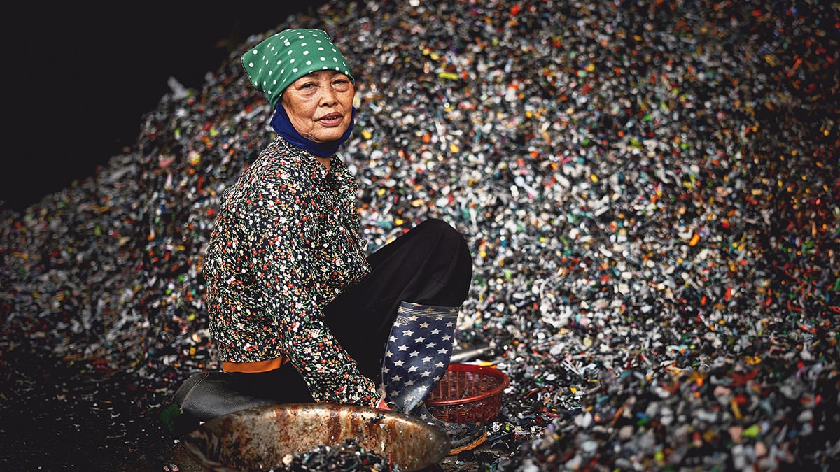 Elderly woman sitting by a large pile of plastic scrap with blue patterned boots and green headscarf, looking at the camera.