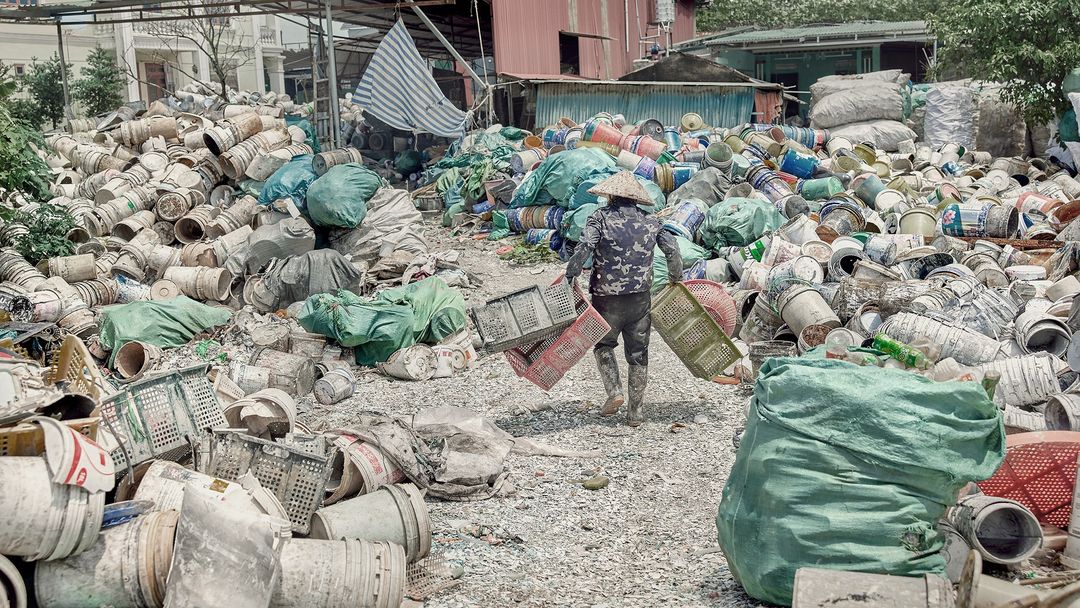 Environmental shot of a person in a conical hat carrying crates through piles of discarded containers in a cluttered outdoor environment.