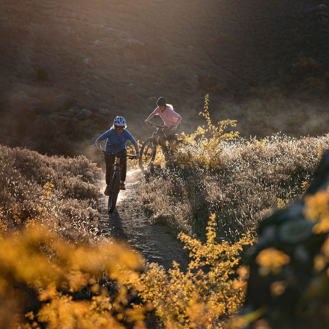 Two women mountain biking on a dirt trail through a grassy, hilly landscape with warm, golden sunlight highlighting the scene.