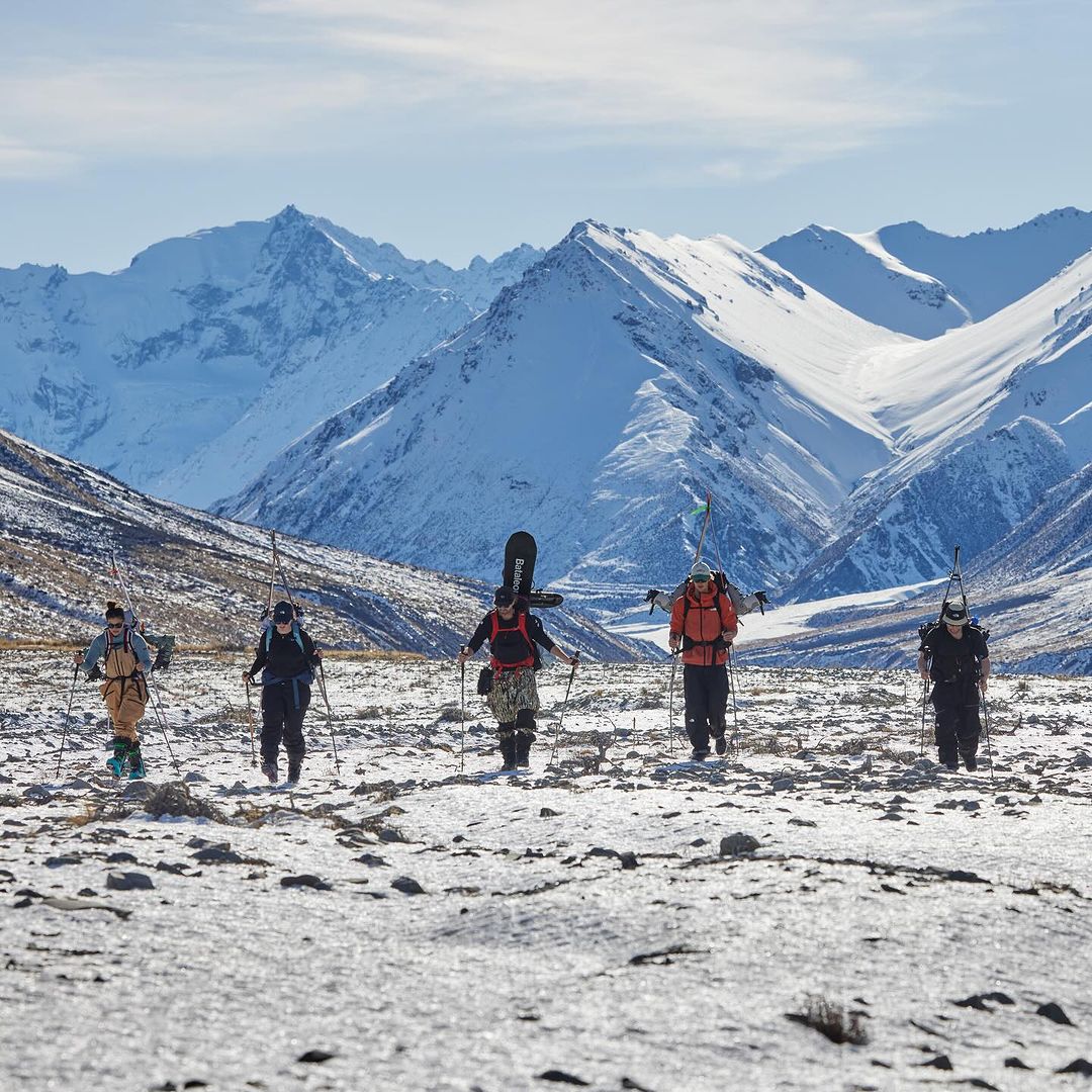 Group of backcountry skiers walking on a snowy plain with skis, poles and snowboards. Majestic snow-capped mountains in the background.