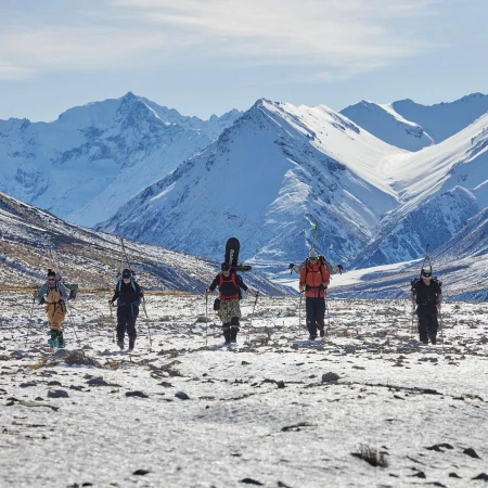 Group of backcountry skiers walking on a snowy plain with skis, poles and snowboards. Majestic snow-capped mountains in the background.