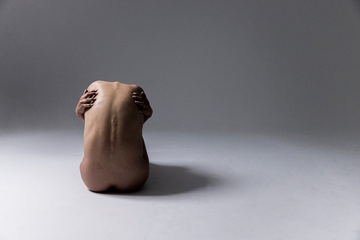 Back view of nude adult female hugging herself while seated on a white surface in a brightly lit, neutral-toned photo studio.