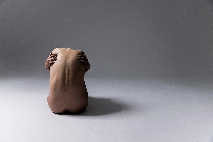 Back view of nude adult female hugging herself while seated on a white surface in a brightly lit, neutral-toned photo studio.