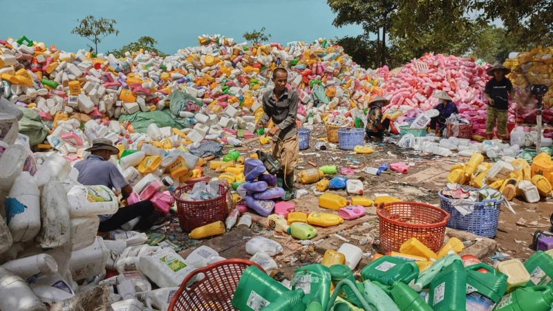 Workers sort through piles of plastic bottles and containers in an outdoor recycling facility, with trees and sky visible in the distance.