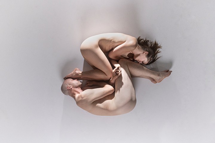 Overhead studio shot of a nude male and female model intertwined on a white backdrop in artistic body arrangement, creating a visually striking composition.
