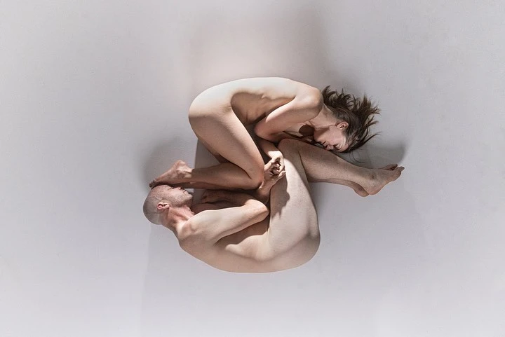 Overhead studio shot of a nude male and female model intertwined on a white backdrop in artistic body arrangement, creating a visually striking composition.