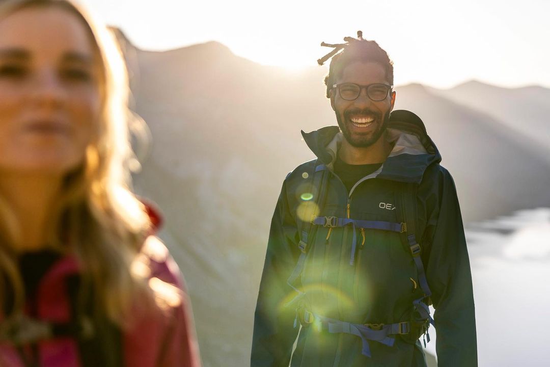 A smiling Black man with glasses and a backpack poses in front of mountains at sunset. A blurred white woman is to his left.