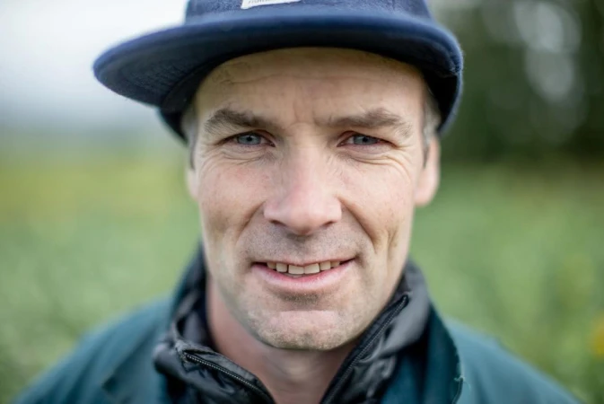 Close-up portrait of a smiling adult man with blue eyes wearing a blue baseball cap outdoors with a blurred green background.