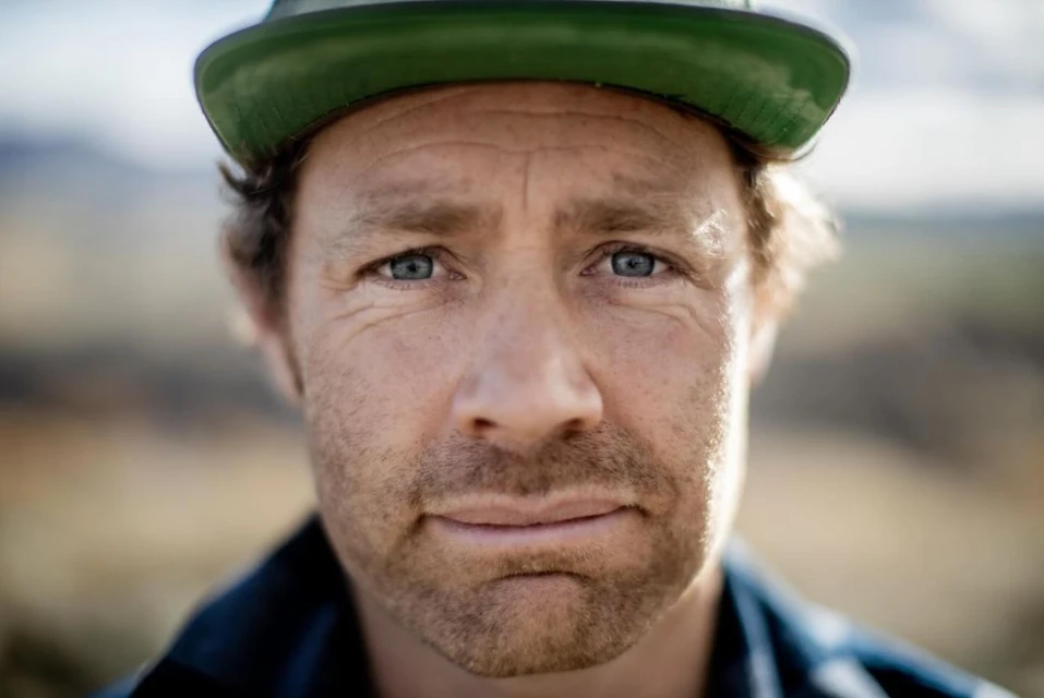 Close-up portrait of an adult male with a green cap, blue eyes, and light stubble, set against a blurred outdoor background.