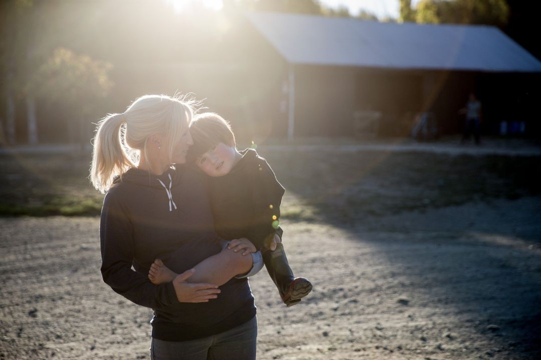 A woman with blonde hair holds a child with a barn and trees in the soft, warm background light of what appears to be golden hour.