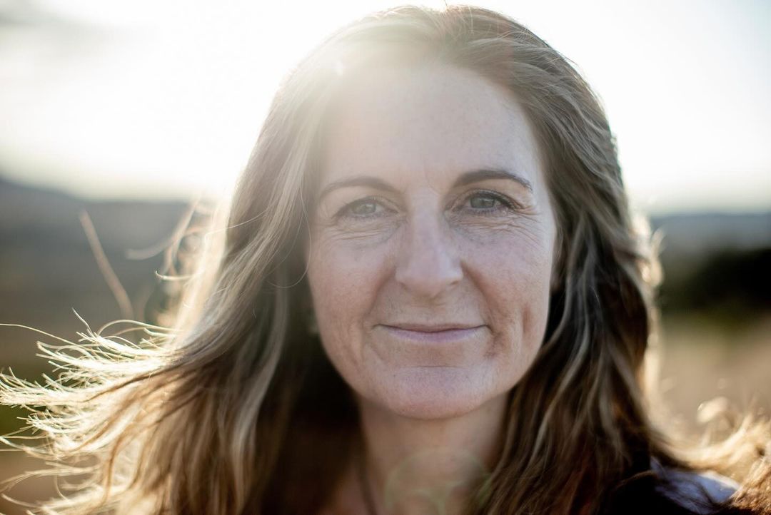 Close-up portrait of an adult woman with shoulder-length hair. She is outdoors with soft, natural lighting and blurred landscape background.