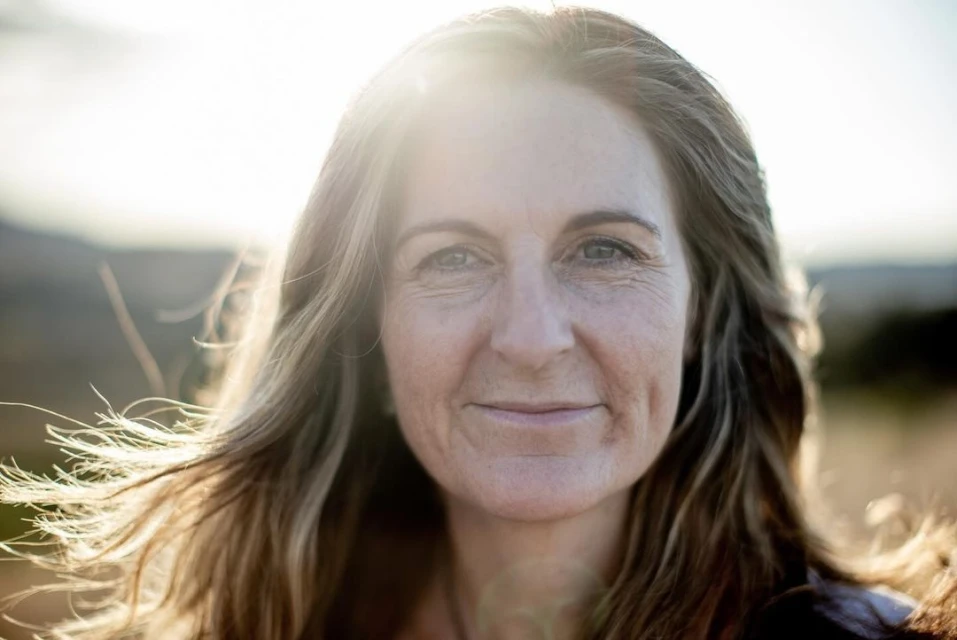 Close-up portrait of an adult woman with shoulder-length hair. She is outdoors with soft, natural lighting and blurred landscape background.