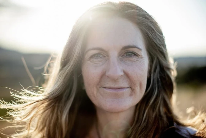 Close-up portrait of an adult woman with shoulder-length hair. She is outdoors with soft, natural lighting and blurred landscape background.