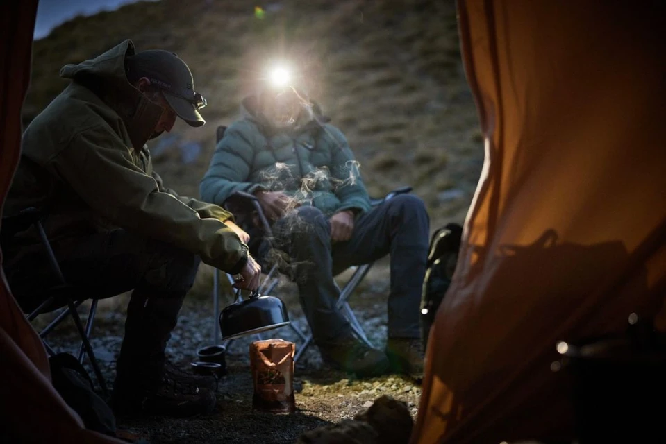 Two men camping outdoors, one pouring from a kettle next to a tent, under a headlamp in a dim, natural outdoor environment.