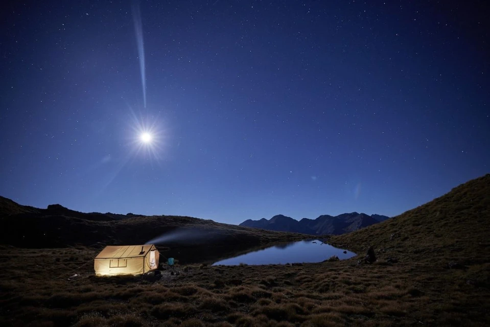 Illuminated tent reflects in alpine lake under a starry night sky with a bright moon and mountain backdrop, creating a serene landscape.