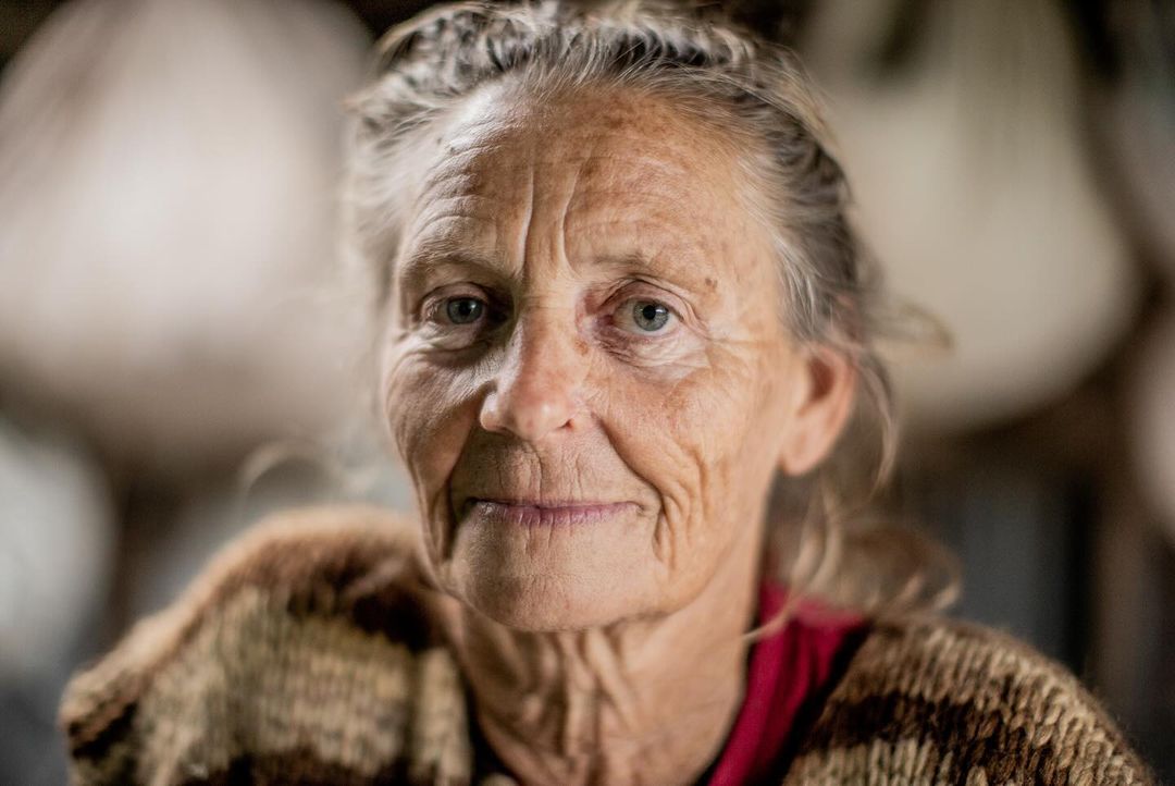 Close-up portrait of a mature woman with fair skin, blue eyes, and gray hair, wearing a striped cardigan, indoors.