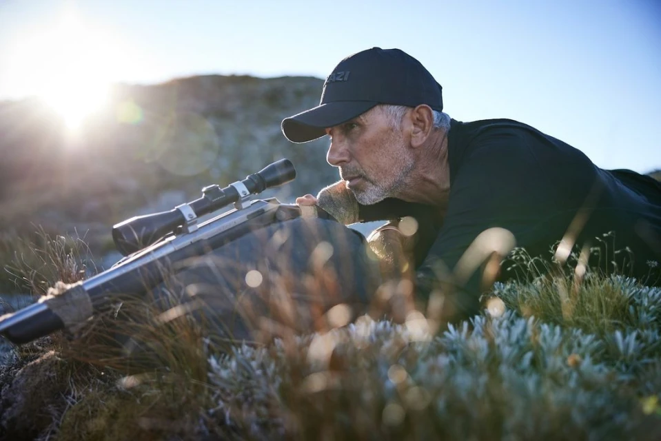 Adult male aiming a rifle with a scope while lying prone on grassy terrain outdoors in bright sunlight, wearing a hat and dark clothing.