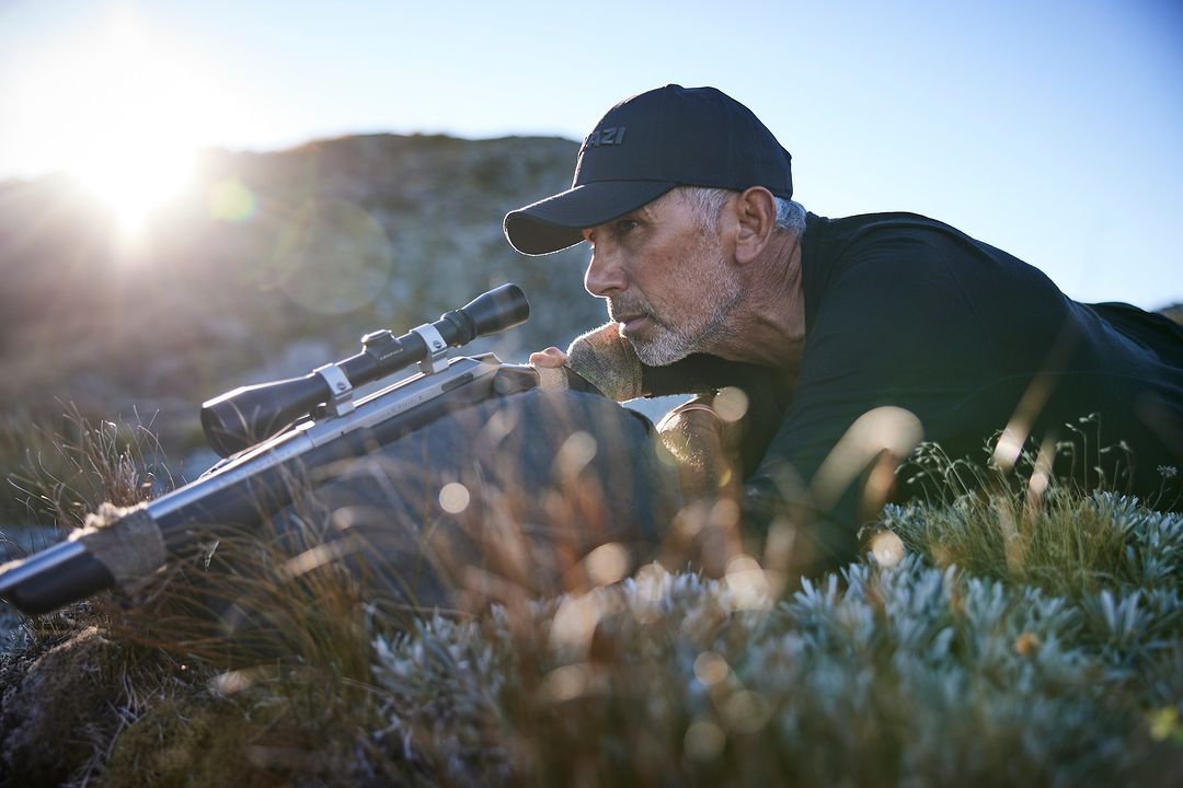 Hunter lying prone in grassy, rocky terrain with a rifle equipped with a scope, aiming towards the sun in the background on a bright day.