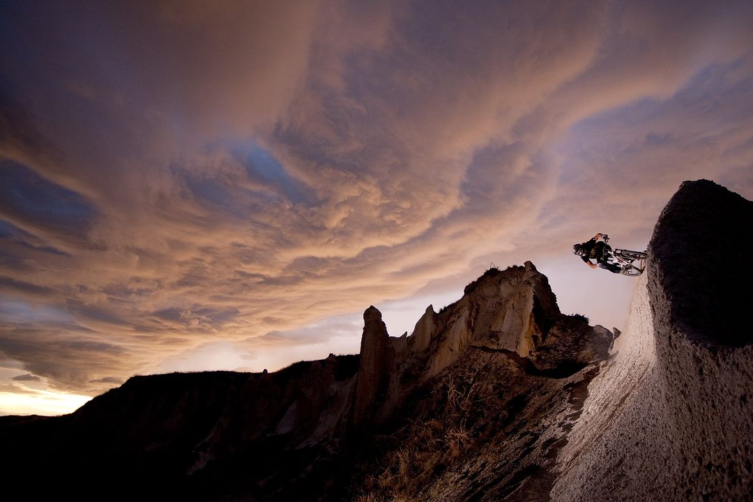 Mountain biker jumps off a rock ledge at sunset, silhouetted against colorful sky. Extreme sports, outdoor adventure in a rocky landscape.