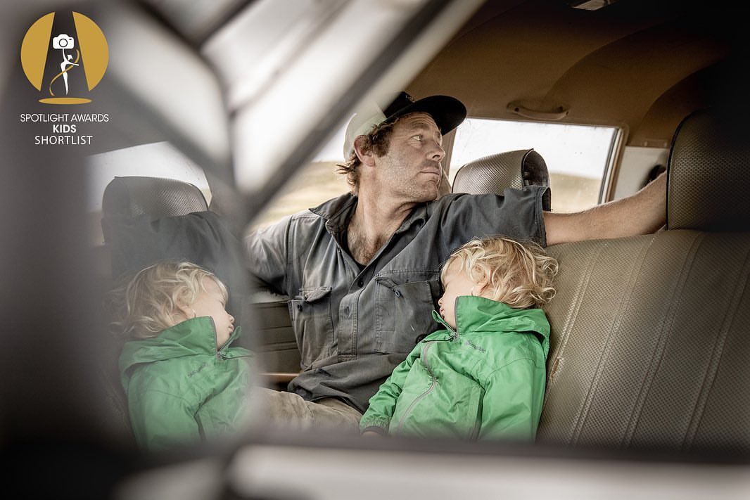 Indoor portrait of a man and two young children seated in a car, featuring natural light and earthy color tones, with a slightly sad mood.