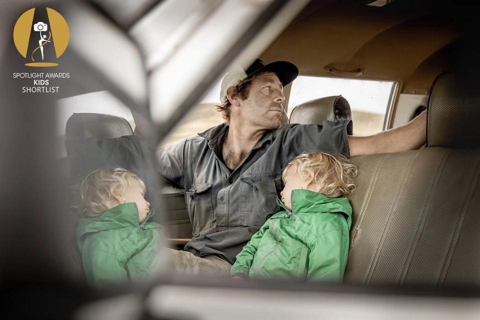 Indoor portrait of a man and two young children seated in a car, featuring natural light and earthy color tones, with a slightly sad mood.
