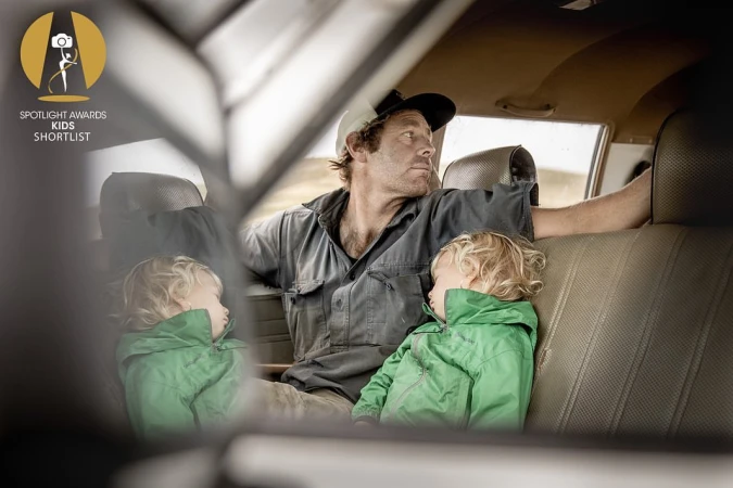 Indoor portrait of a man and two young children seated in a car, featuring natural light and earthy color tones, with a slightly sad mood.